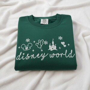 Peut inclure: Un sweat-shirt vert fonc&eacute; pli&eacute;, orn&eacute; de broderies blanches Disney World. Le motif comprend une t&ecirc;te de Mickey Mouse, des ballons, des flocons de neige, un ch&acirc;teau et des c&oelig;urs. Les mots "disney world" sont &eacute;crits en cursive.
