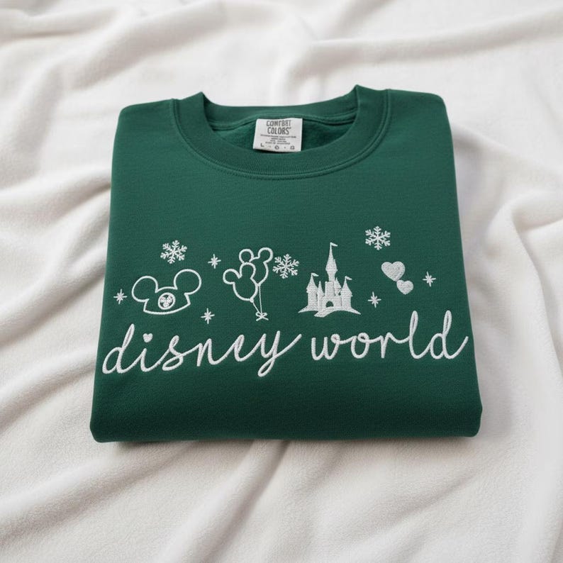 Peut inclure: Un sweat-shirt vert fonc&eacute; pli&eacute;, orn&eacute; de broderies blanches Disney World. Le motif comprend une t&ecirc;te de Mickey Mouse, des ballons, des flocons de neige, un ch&acirc;teau et des c&oelig;urs. Les mots "disney world" sont &eacute;crits en cursive.