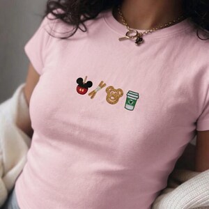 May include: A light pink t-shirt with embroidered Disney-themed icons: a Mickey Mouse apple, churro sticks, a Mickey Mouse-shaped cookie, and a green coffee cup. The shirt has a crew neck and short sleeves.
