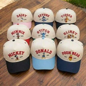 Op de afbeelding: Een verzameling baseballpetten met verschillende Disney-personages. Elke pet heeft een witte kroon, een gekleurde klep en de naam van het personage in het rood geborduurd. Personages zijn Mickey, Minnie, Donald, Daisy, Eeyore, Tigger, Stitch, Slinky en Winnie de Poeh.