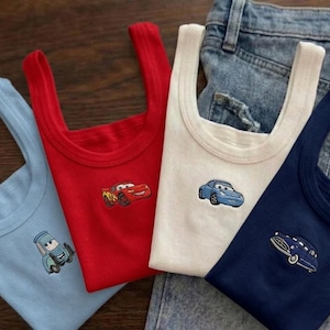 K&ouml;nnte beinhalten: Vier gerippte Tanktops in Hellblau, Rot, Wei&szlig; und Marineblau, jedes mit einem gestickten Cartoon-Auto-Design. Die Tanktops sind gefaltet und in der N&auml;he einer Jeans angeordnet.