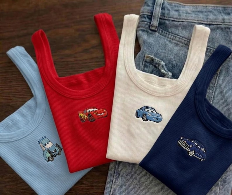 K&ouml;nnte beinhalten: Vier gerippte Tanktops in Hellblau, Rot, Wei&szlig; und Marineblau, jedes mit einem gestickten Cartoon-Auto-Design. Die Tanktops sind gefaltet und in der N&auml;he einer Jeans angeordnet.