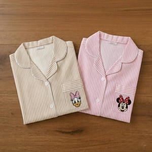 May include: Two pajama tops with a striped pattern. One is beige and the other is pink. Each has a pocket with a cartoon character: Daisy Duck and Minnie Mouse. The pajamas have a button-down front and a collar.
