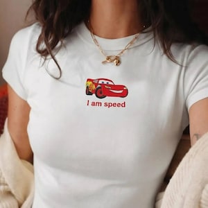 May include: White t-shirt featuring a red cartoon race car with yellow flames and the text "I am speed" in red. The shirt has a crew neck and short sleeves.