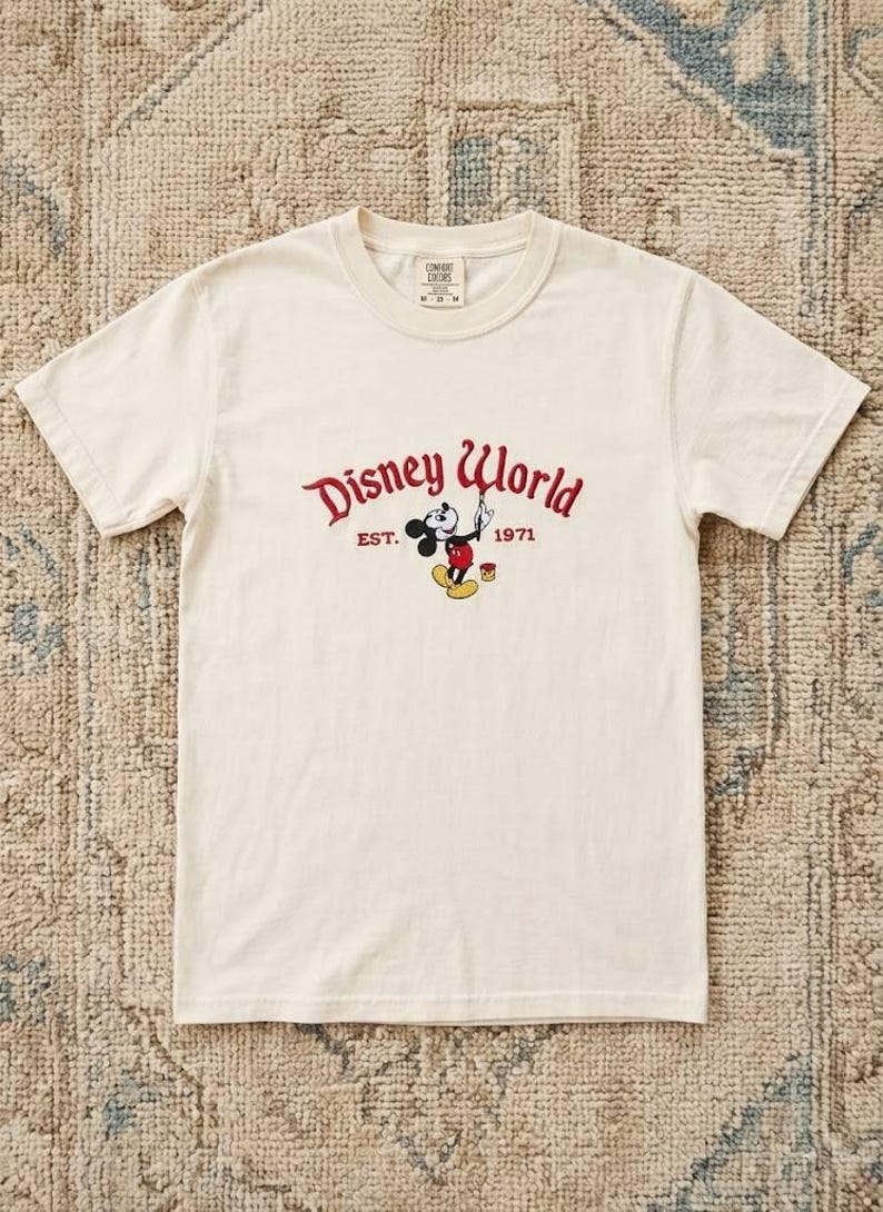 May include: Cream-colored t-shirt featuring the Disney World logo in red with Mickey Mouse. The graphic includes the text "EST. 1971". The shirt has short sleeves and a crew neck.