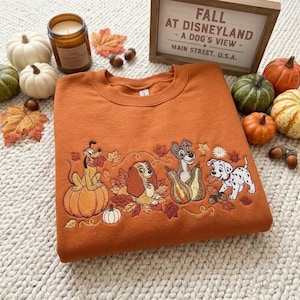 May include: An orange crewneck sweatshirt featuring embroidered Disney dog characters including Pluto, Lady, Tramp, and a Dalmatian puppy surrounded by autumn leaves and pumpkins. The sweatshirt is displayed with a wooden sign reading FALL AT DISNEYLAND.