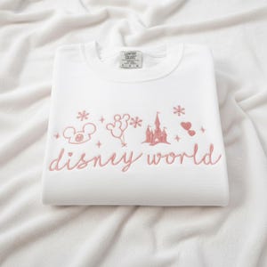 Peut inclure: Sweat-shirt blanc avec broderie rose "disney world" et motifs Disney : Mickey, ballons, ch&acirc;teau, flocons et c&oelig;urs. Le sweat est pli&eacute; sur un fond blanc.