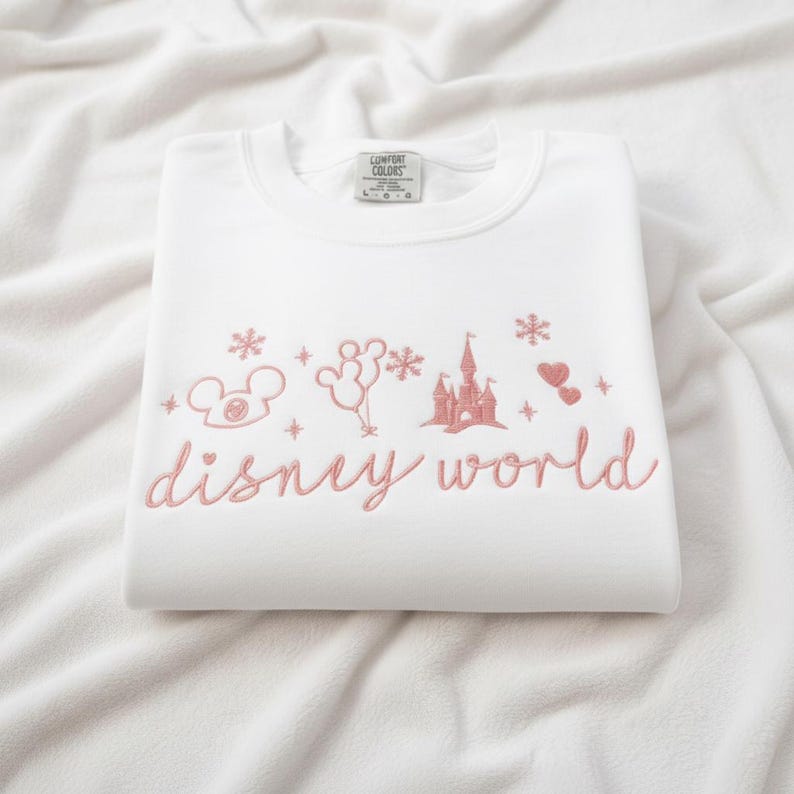 Peut inclure: Sweat-shirt blanc avec broderie rose "disney world" et motifs Disney : Mickey, ballons, ch&acirc;teau, flocons et c&oelig;urs. Le sweat est pli&eacute; sur un fond blanc.