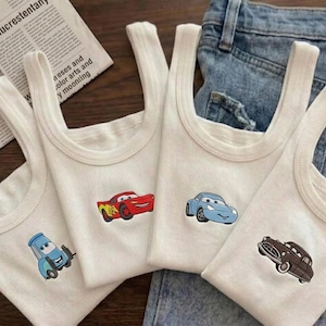K&ouml;nnte beinhalten: Vier wei&szlig;e Ripp-Tanktops mit Cartoon-Autodesigns in Rot, Blau und Braun. Die Tanktops werden auf einem Jeanshintergrund pr&auml;sentiert. Die Autos sind auf die Vorderseite der Tanktops gestickt.