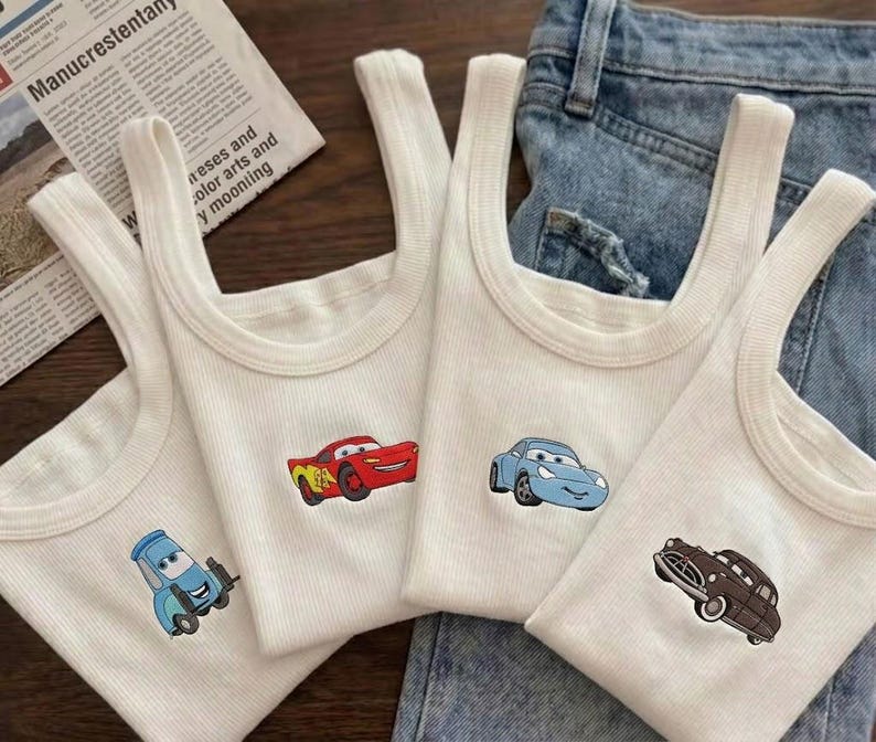 K&ouml;nnte beinhalten: Vier wei&szlig;e Ripp-Tanktops mit Cartoon-Autodesigns in Rot, Blau und Braun. Die Tanktops werden auf einem Jeanshintergrund pr&auml;sentiert. Die Autos sind auf die Vorderseite der Tanktops gestickt.