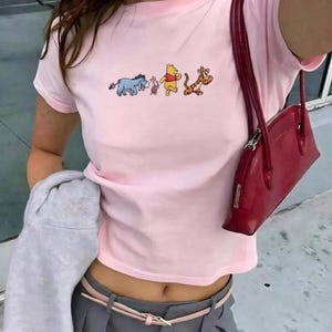 Puede incluir: Camiseta rosa claro con un gráfico de dibujos animados de los personajes de Winnie the Pooh: Ígor, Piglet, Winnie the Pooh y Tigger. La parte superior tiene cuello redondo y mangas cortas.