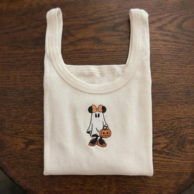 May include: A cream-colored ribbed tank top featuring a central embroidered graphic of a cartoon character dressed as a ghost, wearing an orange polka-dot bow and holding a small orange jack-o'-lantern pumpkin with a carved face.