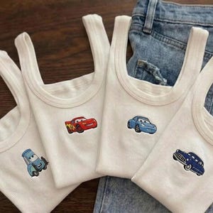 K&ouml;nnte beinhalten: Vier wei&szlig;e Ripp-Tanktops mit gestickten Cartoon-Auto-Designs. Die Autos sind rot, blau und marineblau. Die Tanktops werden auf einer blauen Jeans pr&auml;sentiert.