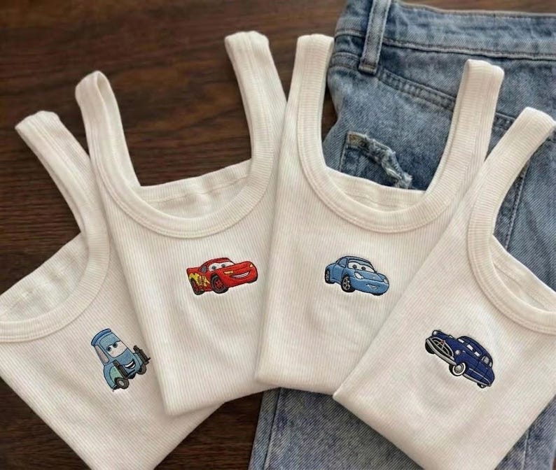 K&ouml;nnte beinhalten: Vier wei&szlig;e Ripp-Tanktops mit gestickten Cartoon-Auto-Designs. Die Autos sind rot, blau und marineblau. Die Tanktops werden auf einer blauen Jeans pr&auml;sentiert.