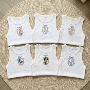 May include: Six white tank tops with embroidered princess portraits. Each top features a different princess in an oval frame, with a bow at the top. The princesses include Rapunzel, Cinderella, and Snow White.