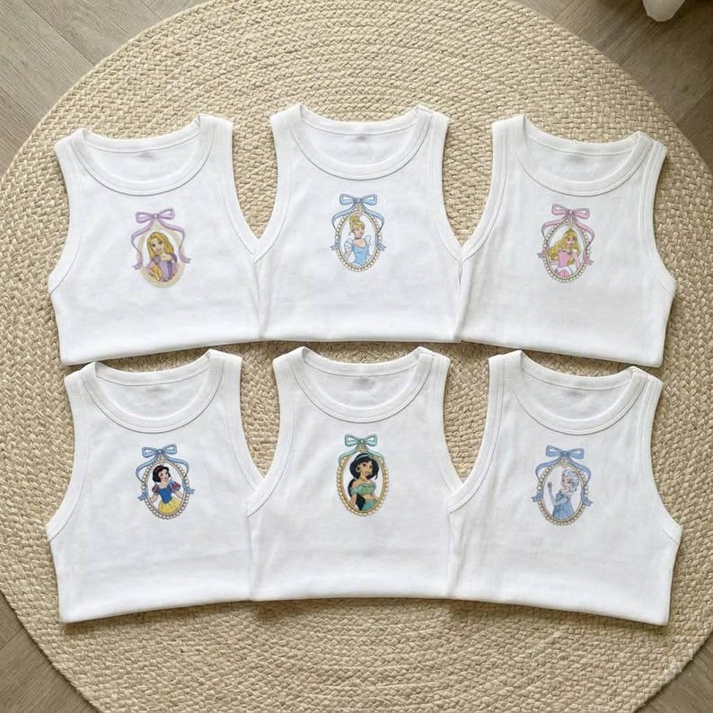 May include: Six white tank tops with embroidered princess portraits. Each top features a different princess in an oval frame, with a bow at the top. The princesses include Rapunzel, Cinderella, and Snow White.