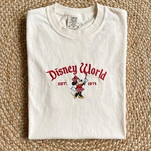 May include: Cream-colored t-shirt featuring the embroidered text "Disney World" in red script, with "EST. 1971" below. A Minnie Mouse graphic is in the center. The shirt is folded on a woven surface.