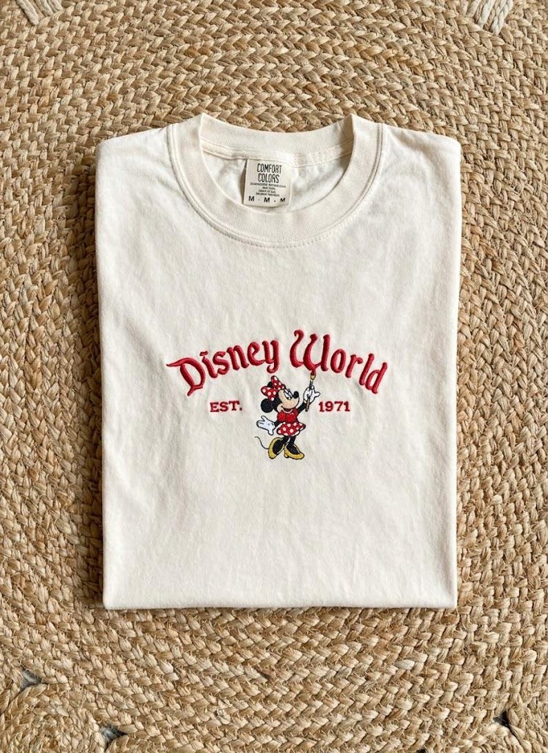 May include: Cream-colored t-shirt featuring the embroidered text "Disney World" in red script, with "EST. 1971" below. A Minnie Mouse graphic is in the center. The shirt is folded on a woven surface.