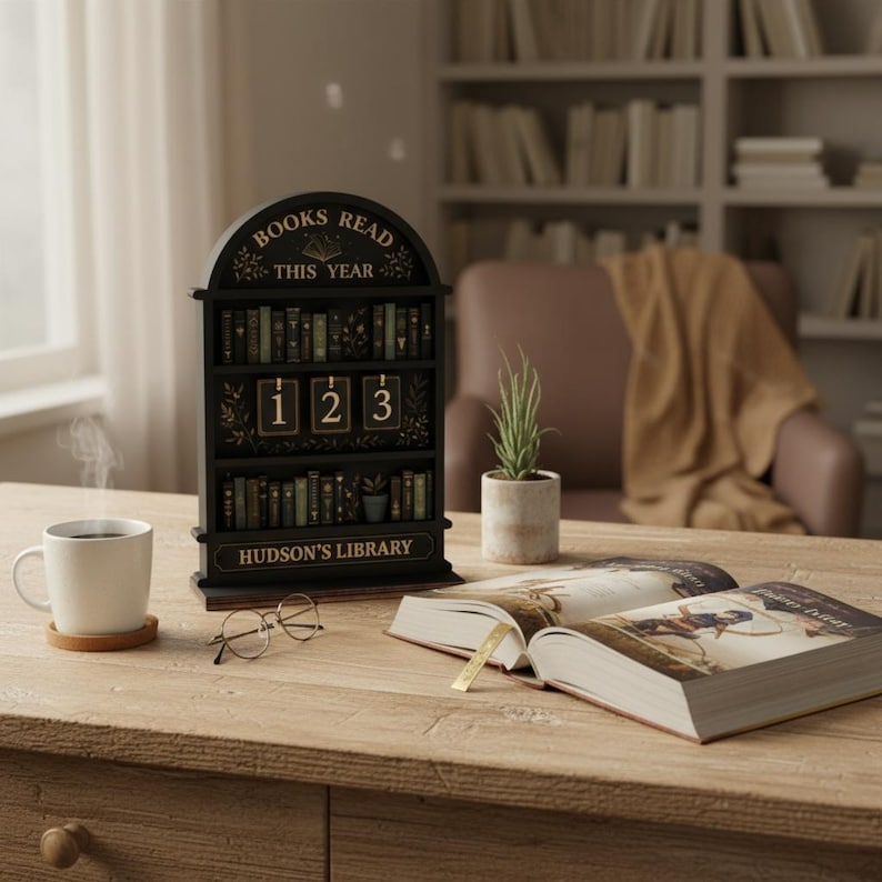 Puede incluir: Un calendario negro en forma de estanter&iacute;a con el texto "BOOKS READ THIS YEAR" y "HUDSON'S LIBRARY". El calendario muestra el n&uacute;mero "123". Una taza blanca, gafas y un libro abierto est&aacute;n sobre una mesa de madera.