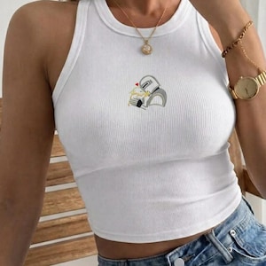 May include: White ribbed tank top with a small embroidered design of two figures embracing. The sleeveless top has a round neckline and is made of a soft, ribbed fabric. The embroidered design features a heart above the figures. The top is paired with blue denim jeans.