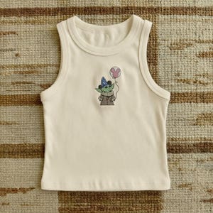 Pu&ograve; includere: Canotta color crema con scollo tondo. La parte anteriore presenta un personaggio dei cartoni animati ricamato con un cappello da festa e un palloncino rosa. Il personaggio &egrave; verde, indossa un cappotto marrone e ha occhi neri.