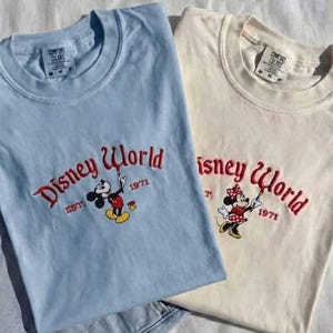 May include: Two t-shirts, one light blue and one cream, featuring the "Disney World" logo in red embroidery. The light blue shirt has Mickey Mouse, and the cream shirt has Minnie Mouse. Both shirts have "EST 1971" below the logo.