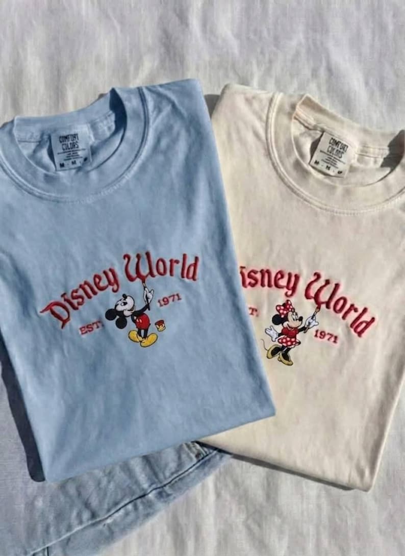 May include: Two t-shirts, one light blue and one cream, featuring the "Disney World" logo in red embroidery. The light blue shirt has Mickey Mouse, and the cream shirt has Minnie Mouse. Both shirts have "EST 1971" below the logo.