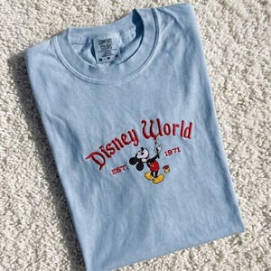 May include: A light blue t-shirt with the words "Disney World" embroidered in red script, along with a Mickey Mouse graphic. The shirt also features the text "EST. 1971". The shirt is folded on a white, textured surface.