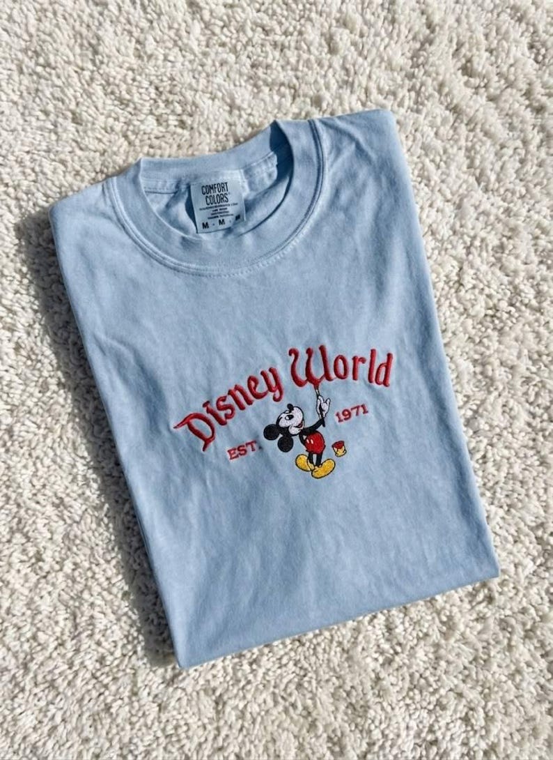 May include: A light blue t-shirt with the words "Disney World" embroidered in red script, along with a Mickey Mouse graphic. The shirt also features the text "EST. 1971". The shirt is folded on a white, textured surface.
