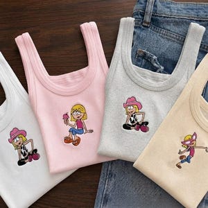 May include: Four ribbed tank tops in white, light pink, heather gray, and cream. Each top features a small embroidered cartoon character graphic on the front. The characters include a blonde figure wearing a pink hat, holding ice cream, or wearing pink sunglasses.