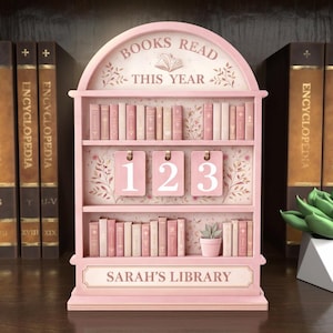 Puede incluir: Un expositor rosa en forma de estanter&iacute;a con el texto "BOOKS READ THIS YEAR" y "SARAH'S LIBRARY". Cuenta con tres estantes con libros en miniatura, tres etiquetas numeradas y una peque&ntilde;a planta en maceta. El fondo incluye enciclopedias.