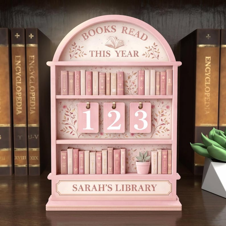 Puede incluir: Un expositor rosa en forma de estanter&iacute;a con el texto "BOOKS READ THIS YEAR" y "SARAH'S LIBRARY". Cuenta con tres estantes con libros en miniatura, tres etiquetas numeradas y una peque&ntilde;a planta en maceta. El fondo incluye enciclopedias.