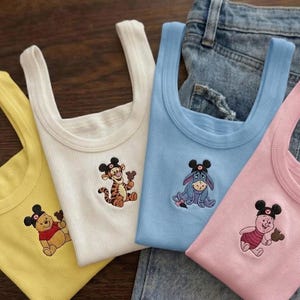 May include: Four ribbed tank tops in yellow, white, light blue, and pink. Each top features an embroidered cartoon character wearing a Mickey Mouse hat. The tops are resting on a pair of blue jeans.