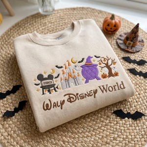 May include: A beige crewneck sweatshirt featuring embroidered Halloween-themed Disney icons, including a castle, a purple cauldron with a witch hat, a tree with pumpkins, and the text Walt Disney World in brown script lettering.
