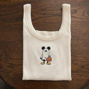 May include: A cream-colored ribbed tank top featuring a central embroidered graphic of a character wearing a white ghost costume. The character has black ears, holds a small orange jack-o'-lantern, and wears yellow shoes, creating a festive look.