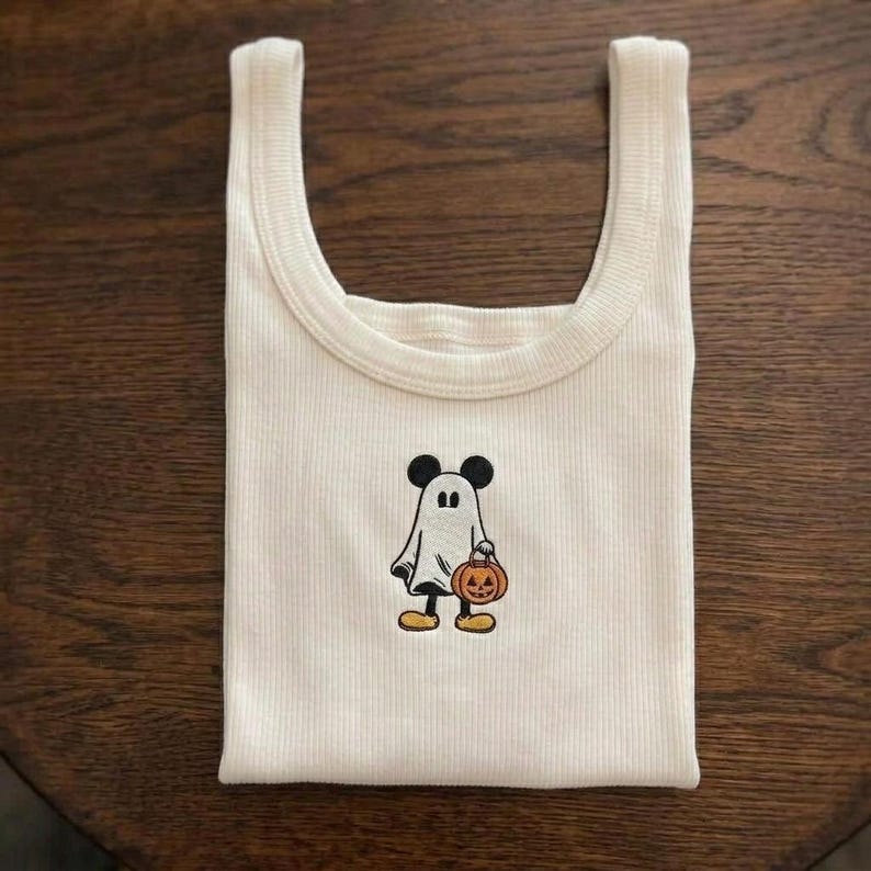 May include: A cream-colored ribbed tank top featuring a central embroidered graphic of a character wearing a white ghost costume. The character has black ears, holds a small orange jack-o'-lantern, and wears yellow shoes, creating a festive look.