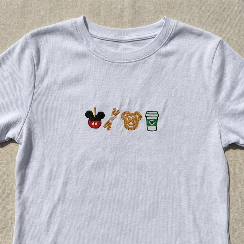 May include: White crew neck t-shirt with embroidered designs of a Mickey Mouse-shaped candy apple, pretzel sticks, a Mickey Mouse-shaped pretzel, and a coffee cup. The embroidery features red, brown, green, and black colors.