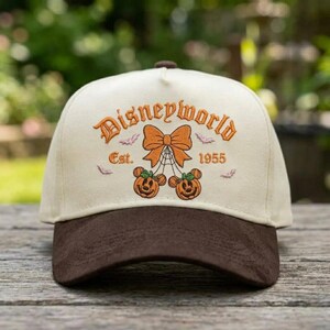 Op de afbeelding: Een crèmekleurige baseballpet met een donkerbruine klep. De voorkant is voorzien van oranje borduursel met de tekst Disneyworld Est. 1955, gecentreerd boven een grote oranje strik, een spinnenweb, twee pompoen-muizenhoofden en kleine paarse vleermuizen.