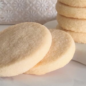 May include: Two round, light brown sugar cookies on a white surface. The cookies are slightly overlapping and have a grainy texture.