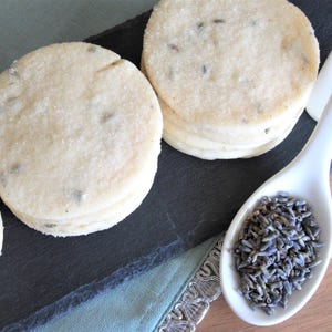 May include: Round, pale beige shortbread cookies with visible dark purple lavender buds embedded in the dough. A white ceramic spoon filled with dried purple lavender buds sits next to the cookies, which are arranged on a dark gray slate board.