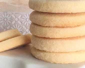 Può includere: Una pila di cinque biscotti rotondi di pasta frolla color beige chiaro con una superficie granulosa e strutturata. Due biscotti aggiuntivi sono appoggiati in piano sulla superficie bianca accanto alla pila. Hanno spessore uniforme e colore dorato.