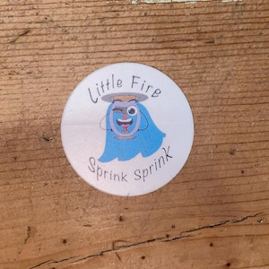 Peut inclure: Un autocollant rond blanc avec le texte "Little Fire" en police incurvée. L'autocollant présente un personnage de dessin animé avec un corps bleu, un visage souriant et un halo. Les mots "Sprink sprink" sont écrits en dessous.