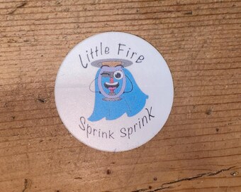 Puede incluir: Una pegatina redonda blanca con el texto "Little Fire" en una fuente curva. La pegatina presenta un personaje de dibujos animados con un cuerpo azul, una cara sonriente y un halo. Las palabras "Sprink sprink" están escritas debajo.