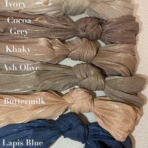 Puede incluir: Colección de coleteros de tela en varios colores, incluyendo marfil, cacao, gris, caqui, verde oliva ceniza, suero de mantequilla y azul lapislázuli. Cada coletero está anudado y muestra un aspecto brillante y texturizado. Los nombres de los colores están impresos.