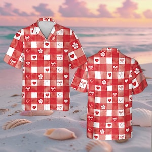 May include: Red and white checkered short-sleeve shirt with a repeating pattern of hearts, bows, and flowers. The shirt has a classic collar and is displayed on a sandy beach setting.