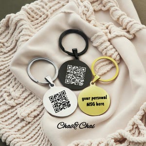 May include: Three round keychains in silver, black, and gold. The silver and black keychains have a QR code, while the gold keychain has the text "your personal MSG here". The keychains are on a beige fabric.