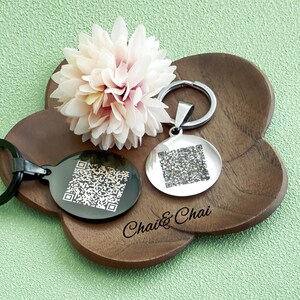 May include: Two round keyrings with QR codes, one black and one silver, sit on a wooden tray with a flower. The black keyring has a black ring, and the silver keyring has a silver ring. The tray has the text "Chai & Chai".