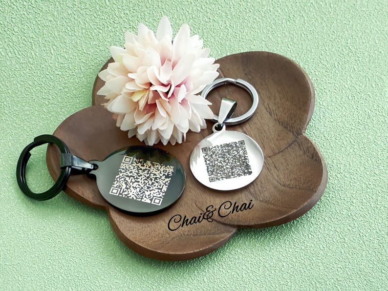 May include: Two round keyrings with QR codes, one black and one silver, sit on a wooden tray with a flower. The black keyring has a black ring, and the silver keyring has a silver ring. The tray has the text "Chai & Chai".