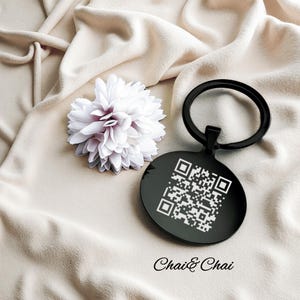 May include: A black round keychain with a QR code, next to a white flower with purple accents. The keychain has a black ring and the text "Chai & Chai" is at the bottom.