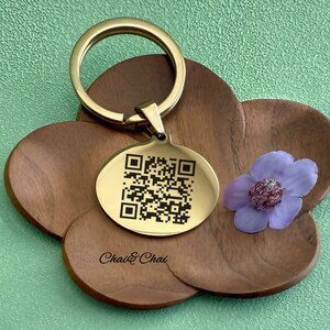 May include: A gold-coloured keyring with a round tag featuring a QR code. The keyring is attached to a wooden, flower-shaped coaster. The text "Chai & Chai" is printed on the coaster. A purple flower with a dark centre is also present.